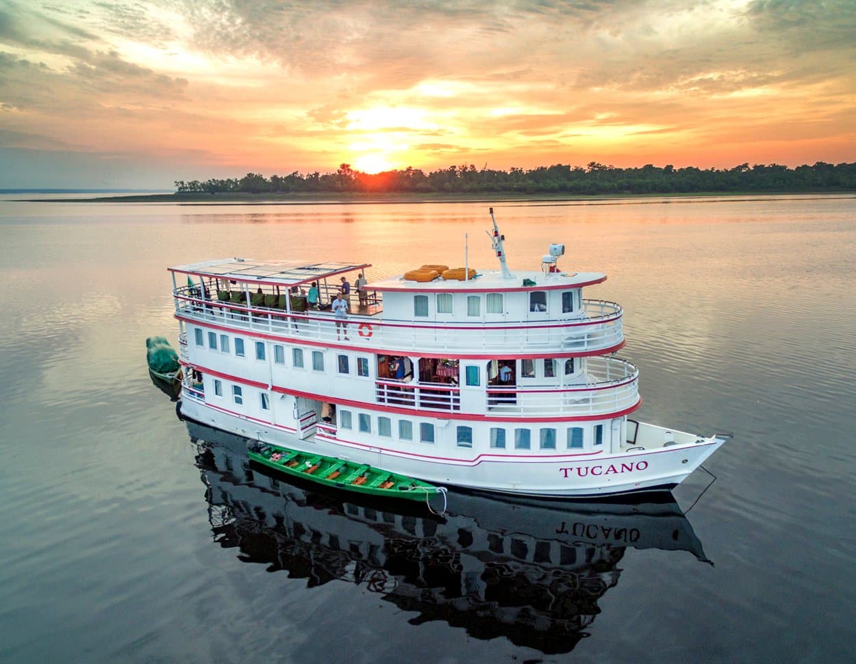 Fünf Nächte an Bord, und der Abend gehört dem Fluss. Das M/Y Tucano, ein weißes Flusskreuzfahrtschiff mit drei Decks, liegt bei Sonnenuntergang auf einem spiegelglatten Schwarzwasserfluss, ein grünes Holzkanu liegt längsseits, Passagiere stehen auf dem Oberdeck, Waldsilhouette vor orangefarbenem Himmel