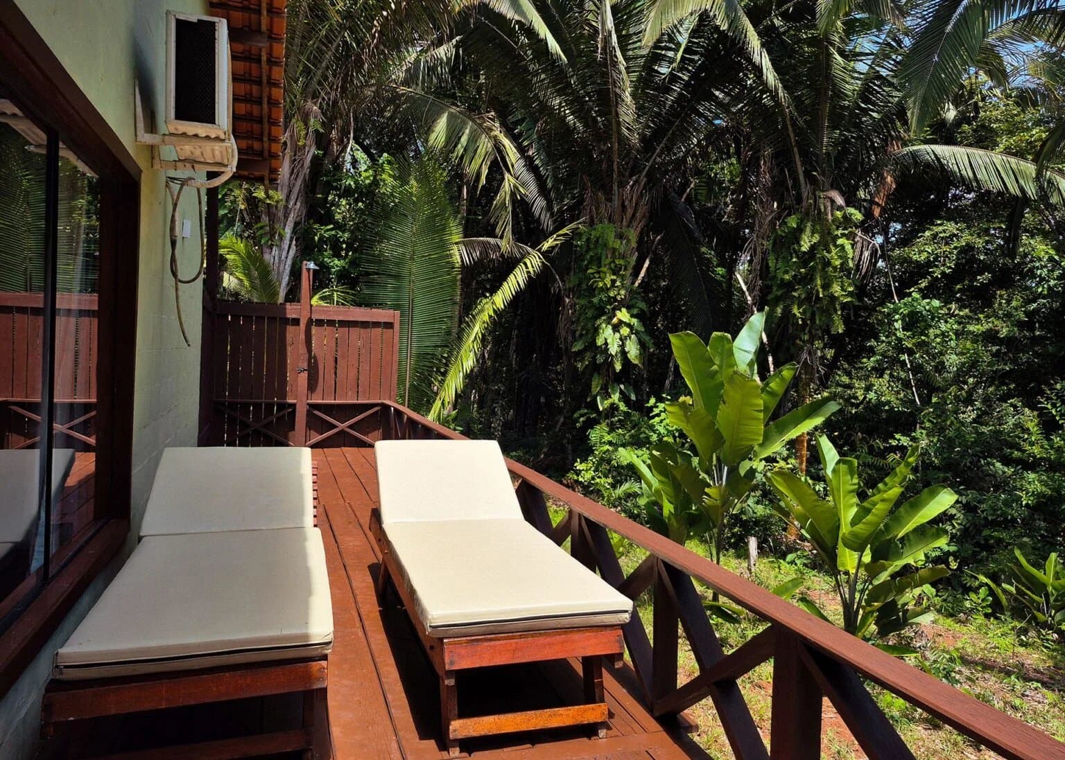 Zwei Stühle, kein Plan, und der Regenwald macht einfach weiter. Zwei gepolsterte Holzliegen auf einer privaten Holzterrasse einer Amazon Boutique Lodge, umgeben von dichter tropischer Vegetation mit Palmen und Bananenstauden