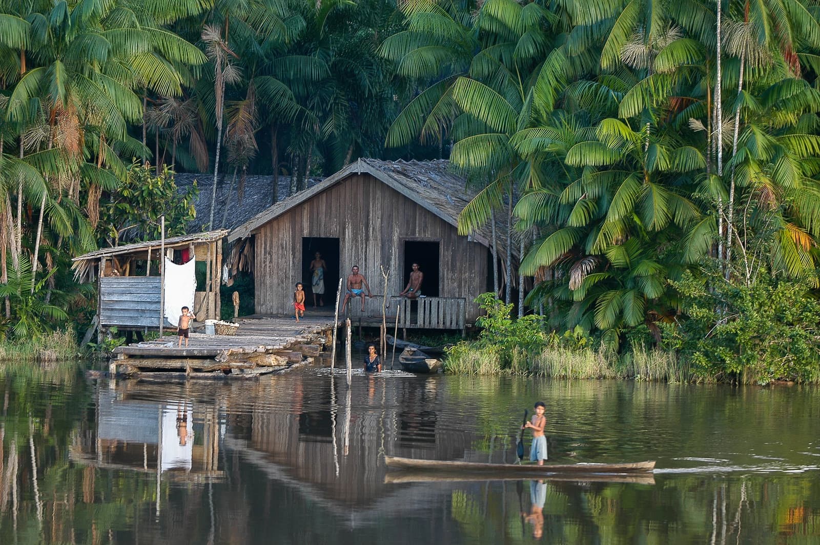 Kein Fahrplan, kein Lärm, nur der Fluss und sein Rhythmus. Ein Holzhaus auf Stelzen am Amazonas-Ufer, umgeben von dichtem Palmenwald, mit einer Familie auf der Veranda und dem Steg, einer Person, die im seichten Wasser steht, und einem Kind in einem Einbaum auf dem stillen Fluss — Einblick in das Leben der Caboclo-Gemeinden auf einer Amazonas-Kreuzfahrt in Brasilien