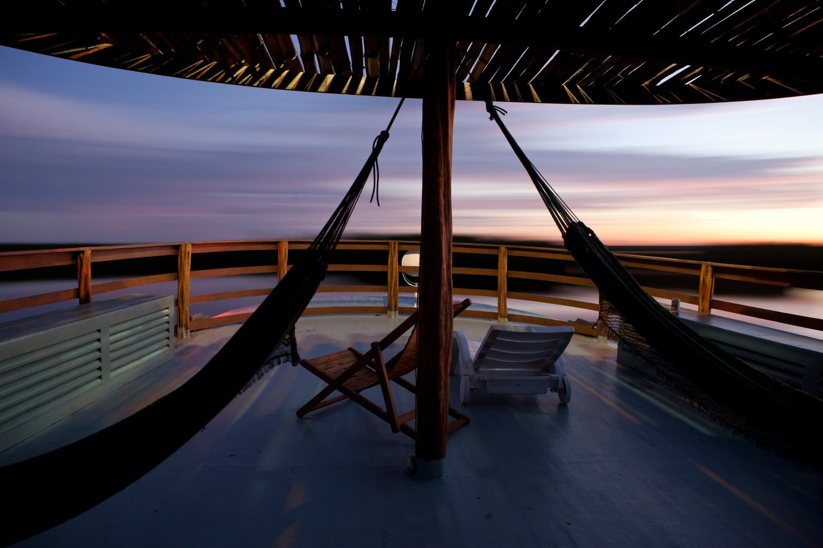 Zwei Hängematten, ein Fluss und kein Grund zur Eile. Offenes Oberdeck eines Amazonas-Flusskreuzfahrtschiffs in der Abenddämmerung, mit zwei Hängematten an einem Palapa-Mast, Holzliegestühlen und einem weiten Blick auf den orangefarben und lila leuchtenden Horizont über dem Fluss