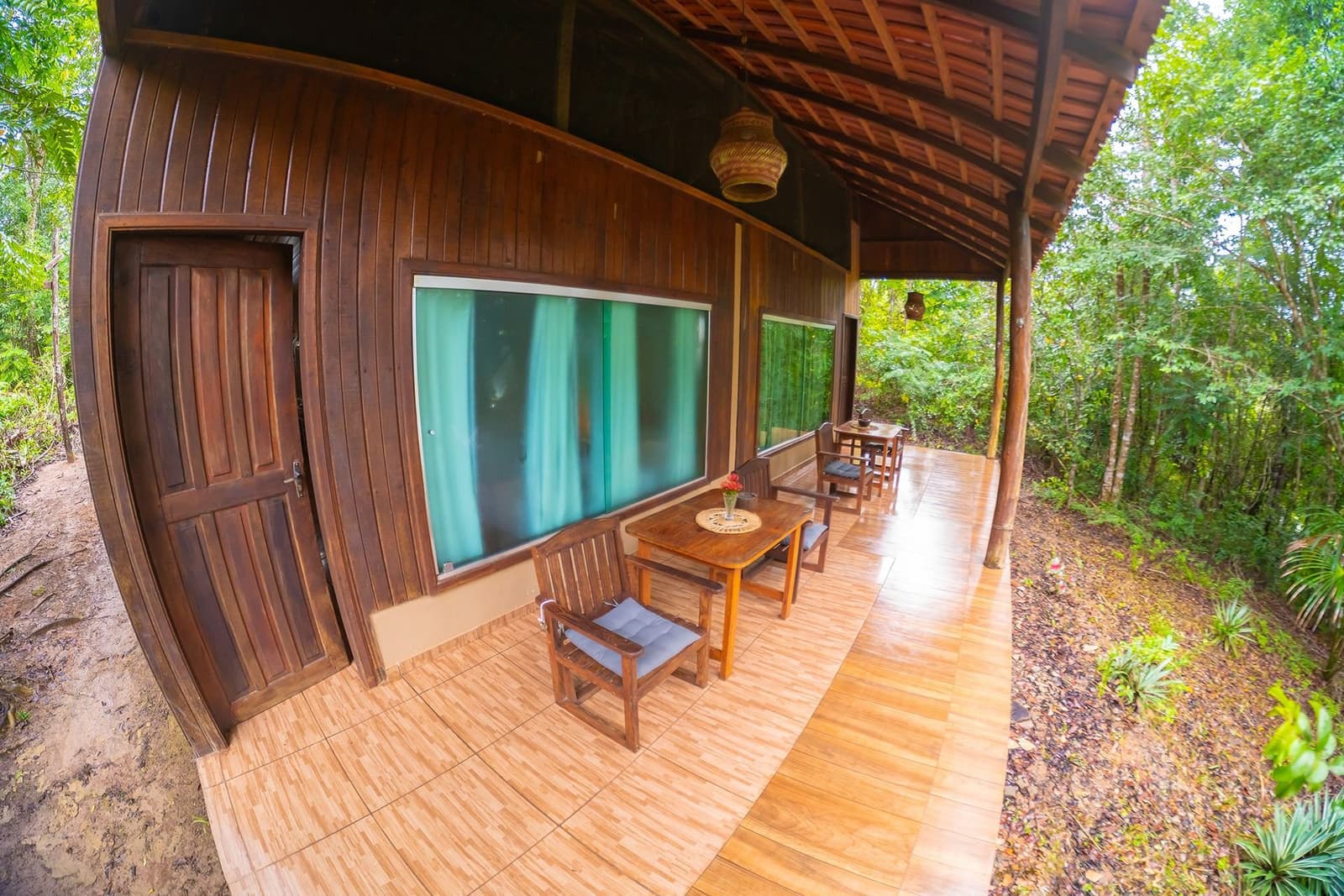 Regenwald direkt vor der Tür, Frühstückskaffee inklusive. Überdachte Holzveranda der Amazon Turtle Lodge mit Holzstühlen, gepolsterten Sitzen und kleinen Esstischen entlang eines Bungalows aus dunklem Holz, umgeben von dichtem Regenwald