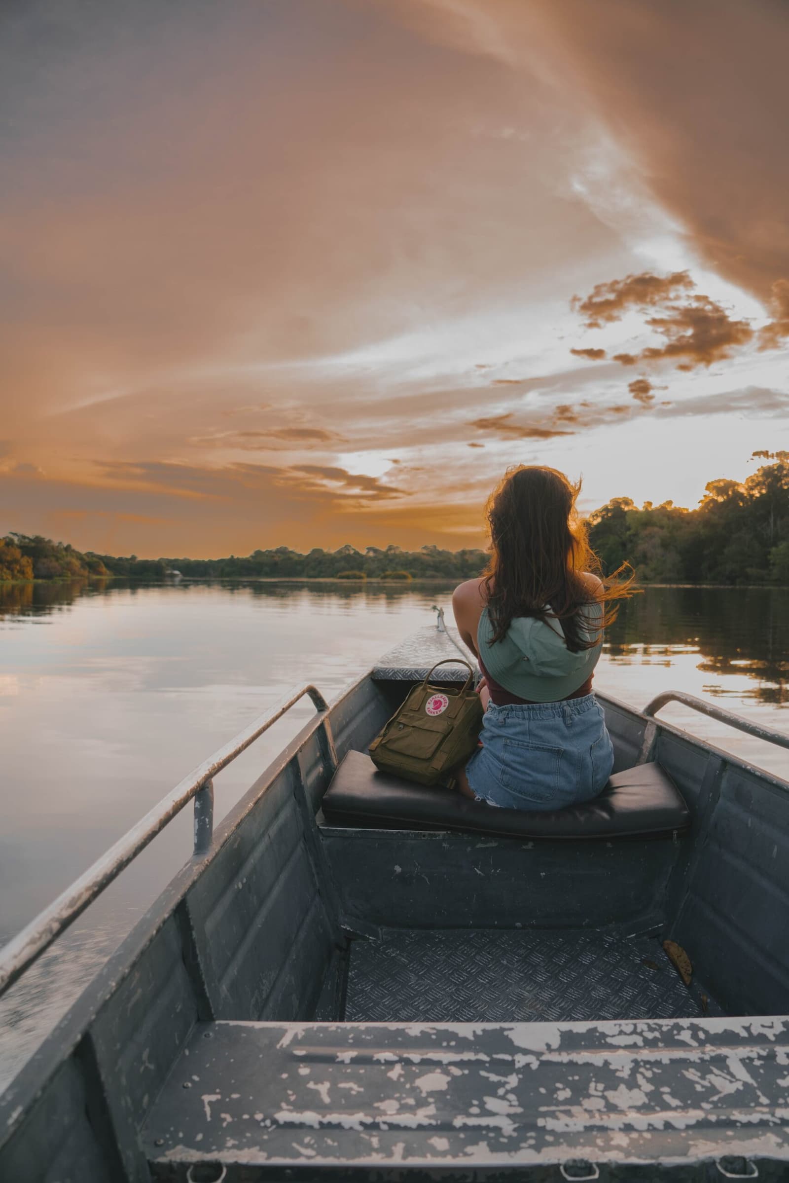 Manche Momente brauchen kein Ziel, nur den Fluss und das Licht. Eine Reisende sitzt am Bug eines Flussbootes und betrachtet einen leuchtend orangefarbenen Sonnenuntergang über einem ruhigen, von Dschungel gesäumten Fluss beim Amazon Turtle Lodge