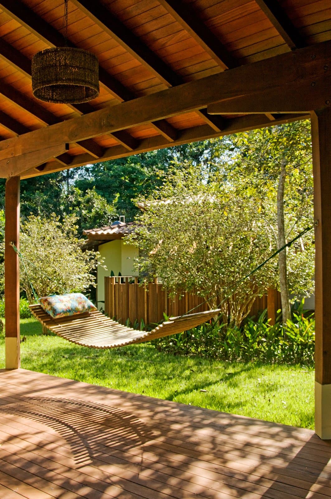 Entre deux sorties, le hamac vous attend, et c'est une très bonne raison de rentrer déjeuner. Photo: Luis Gomes Un hamac en lattes de bois avec un coussin fleuri suspendu sous une pergola à poutres apparentes sur une terrasse en bois, encadrant un jardin verdoyant et un petit bâtiment de lodge, dans un lodge d'observation des oiseaux en forêt amazonienne