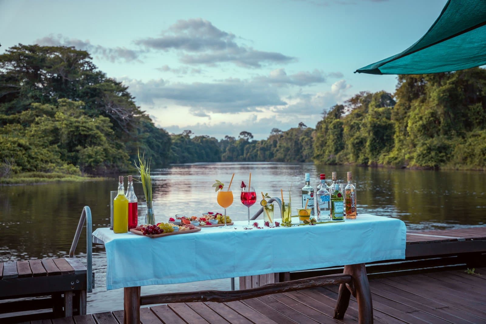 L'heure dorée sur le ponton, le fleuve en pause, la forêt à portée de verre. Photo: Samuel Melim Une table drapée de blanc sur une terrasse en bois au bord de l'eau, garnie de cocktails tropicaux, de planches de fruits et fromages et de bouteilles de spiritueux, avec la forêt amazonienne dense bordant un canal fluvial calme au crépuscule