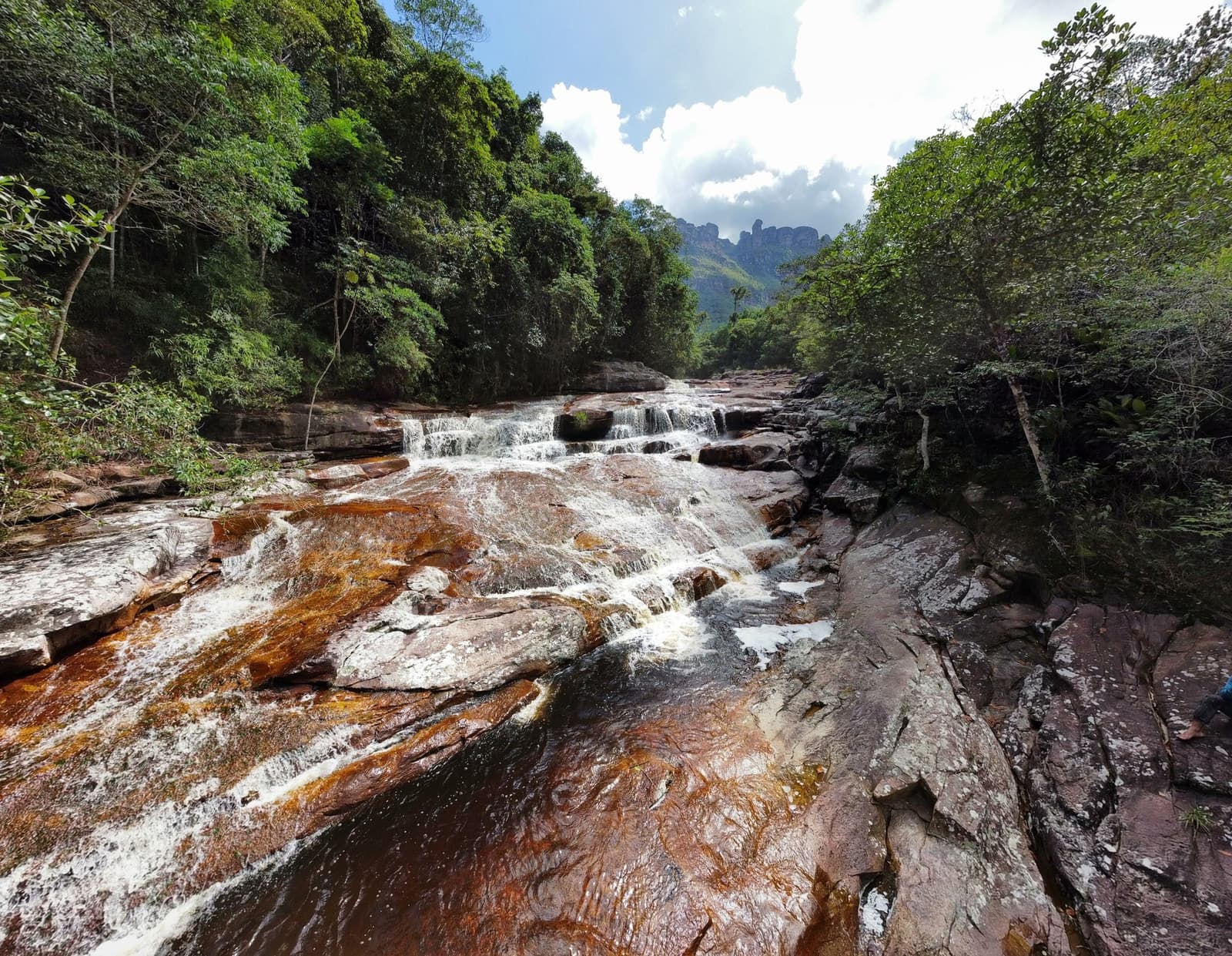 Eisenrotes Gestein, kühles Wasser, nackte Füße. Der Pati-Fluss macht, was er will. Klares Wasser strömt über rostfarbene Sandsteinabsätze in einer gestuften Kaskade beim Chapada-Diamantina-Trek, mit dichtem Regenwald und zerklüfteten Felsformationen im Hintergrund