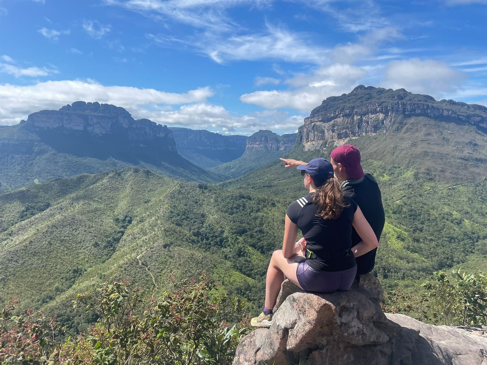 Einer zeigt immer genau auf den richtigen Gipfel zuerst. Photo: Lucas Neves Zwei Wanderer sitzen auf einem Felsen an einem Aussichtspunkt beim Chapada-Diamantina-Trek und zeigen auf ein weitläufiges grünes Tal, das von mächtigen Sandstein-Tafelbergen gesäumt wird