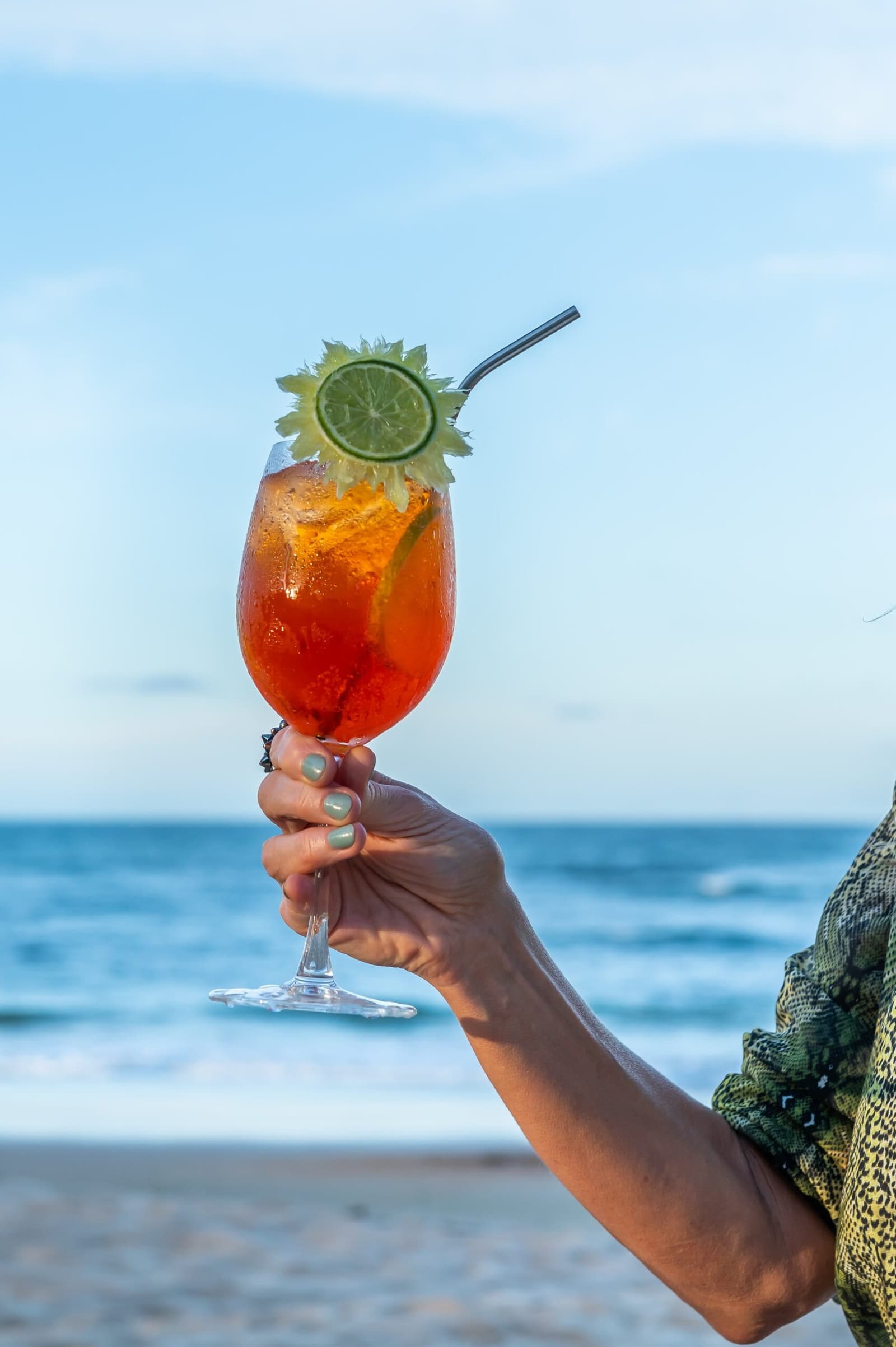 Verdient. Ein kaltes Glas, der Ozean, mehr braucht es nicht. Eine Hand hält ein großes, eisgekühltes rotes Cocktailglas mit Limettenscheibe und Kräutern, im Hintergrund das Meer an der Küste von Bahia
