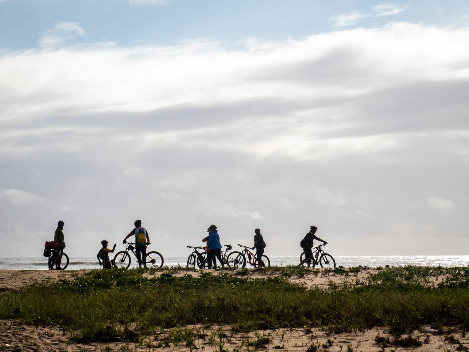 Sechs Räder, ein Horizont, und der beste Teil kommt noch. Gruppe von Radfahrern mit Mountainbikes, die auf einer sandigen Küstendüne mit niedrigem Bewuchs pausieren, als Silhouette gegen einen hellen bewölkten Himmel auf der Bahia-Radroute