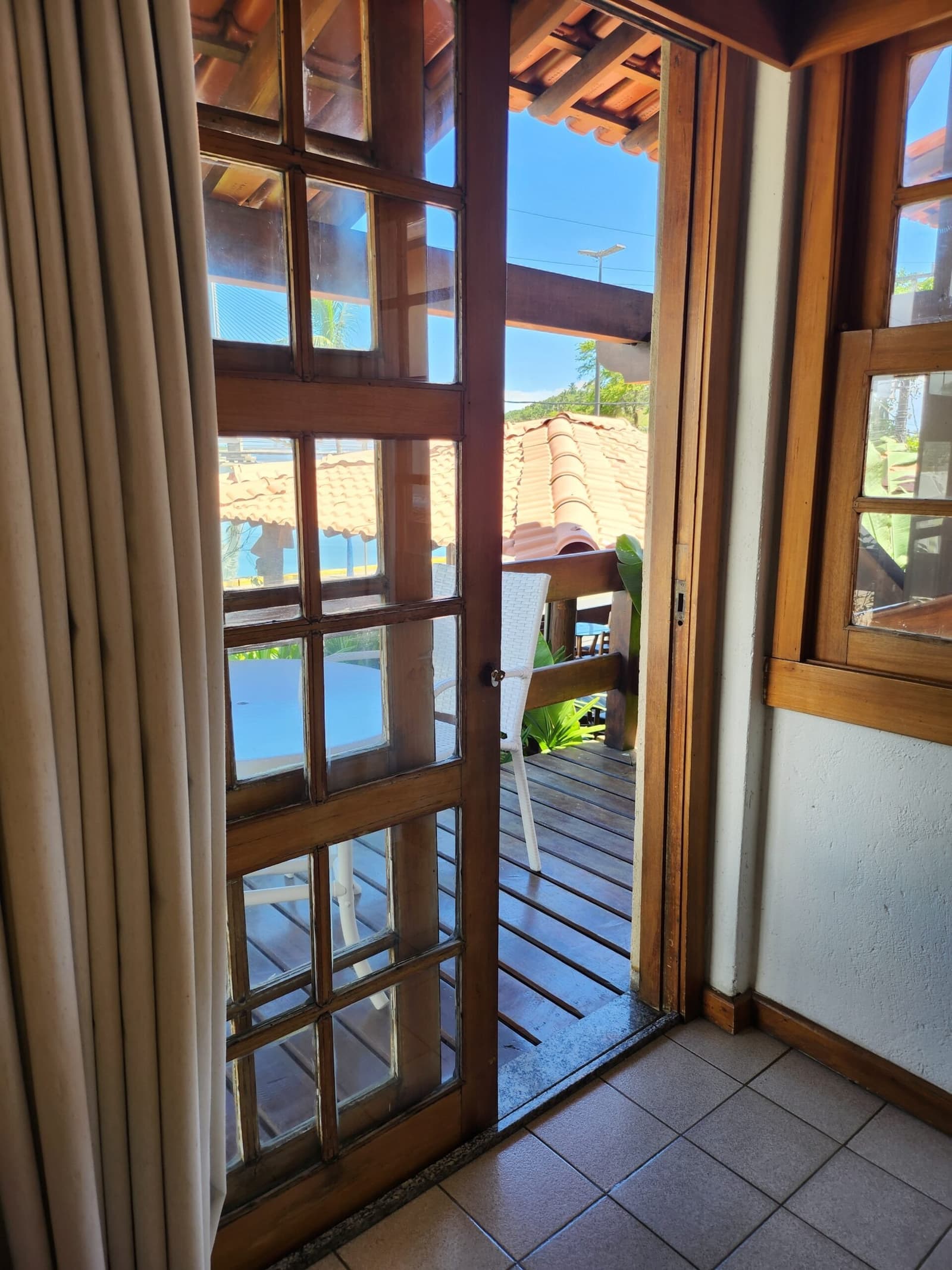 Die Tür steht offen, der Liegestuhl auch. Einfach hingehen. Blick durch eine geöffnete Holzrahmen-Glastür auf eine sonnenbeschienene Holzterrasse mit weißem Stuhl, Terrakottadach und tropischem Grün in einer Pension