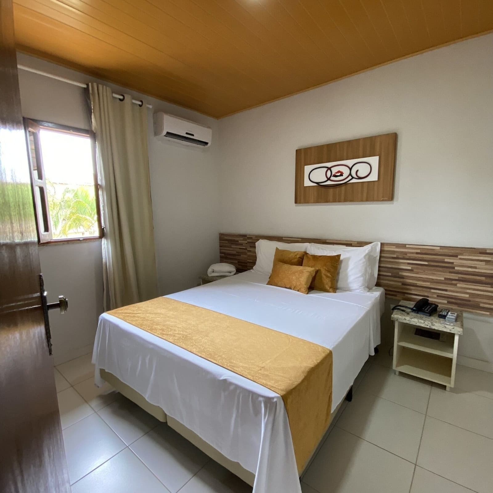 Frische Bettwäsche, kühle Luft, gute Nacht. Sauber gemachtes Doppelbett mit goldenen Zierkissen und Bettläufer in einem klimatisierten Hotelzimmer mit Holzdecke und Blick auf tropisches Grün, Unterkunft auf einer Radtour durch Bahia