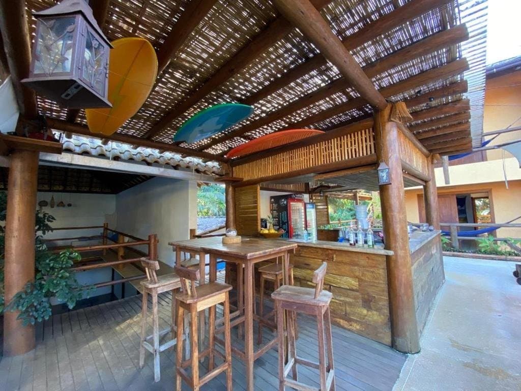 Hocker frei, Getränke kalt, Kajaks an der Decke, hier stimmen die Prioritäten. Offene Holzbar mit Baumstamm-Pergola, Bambusdecke, Barhockern und bunten Kajaks an der Decke in einer Unterkunft auf der Bahia-Radroute