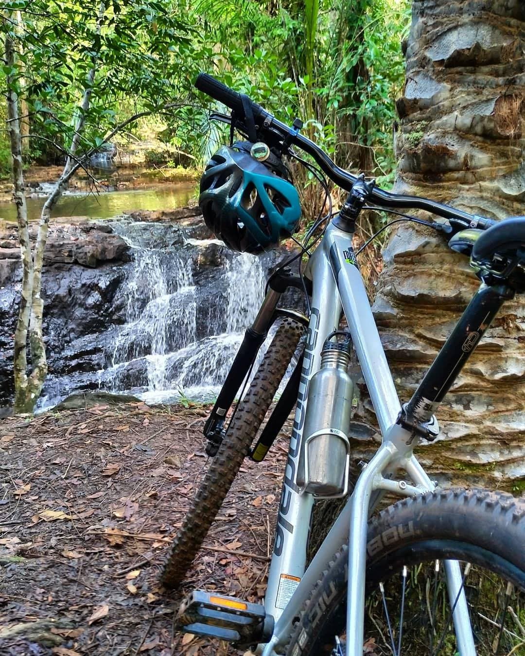Manche Pausen braucht es einfach. Rad ab, Helm ab, Wasserfall an. Ein Mountainbike mit türkisfarbenem Helm lehnt an einem Sandsteinfelsenblock neben einem kleinen Wasserfall auf einem Radweg durch den Atlantischen Regenwald in Bahia