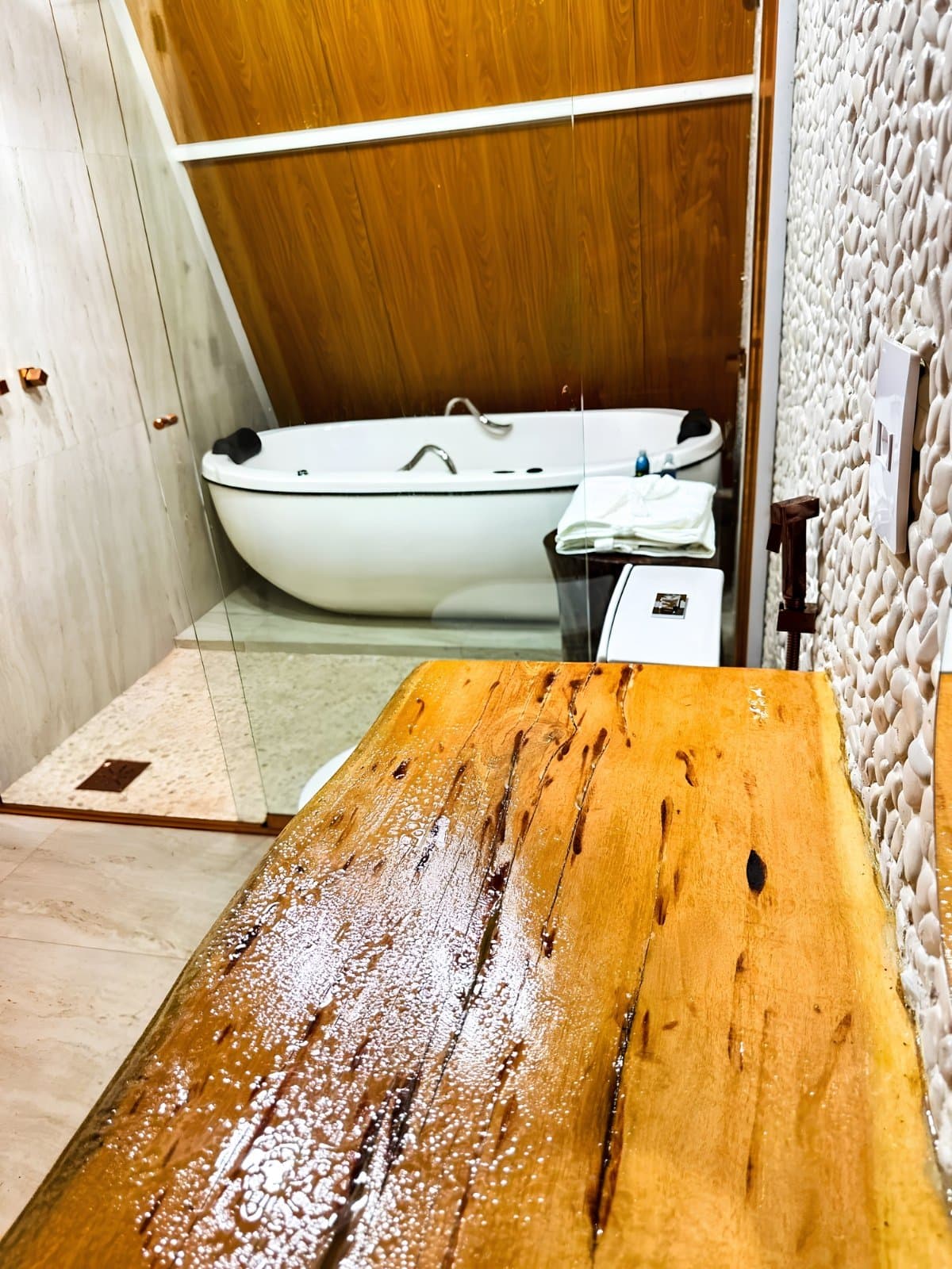 Trilha, fervedouro, poeira, e no fim: isso aqui. Justo. Uma banheira oval branca encostada a paredes de madeira aquecida em um banheiro de pousada, com um banco de madeira bruta molhado em primeiro plano e uma parede de pedra seixo texturizada