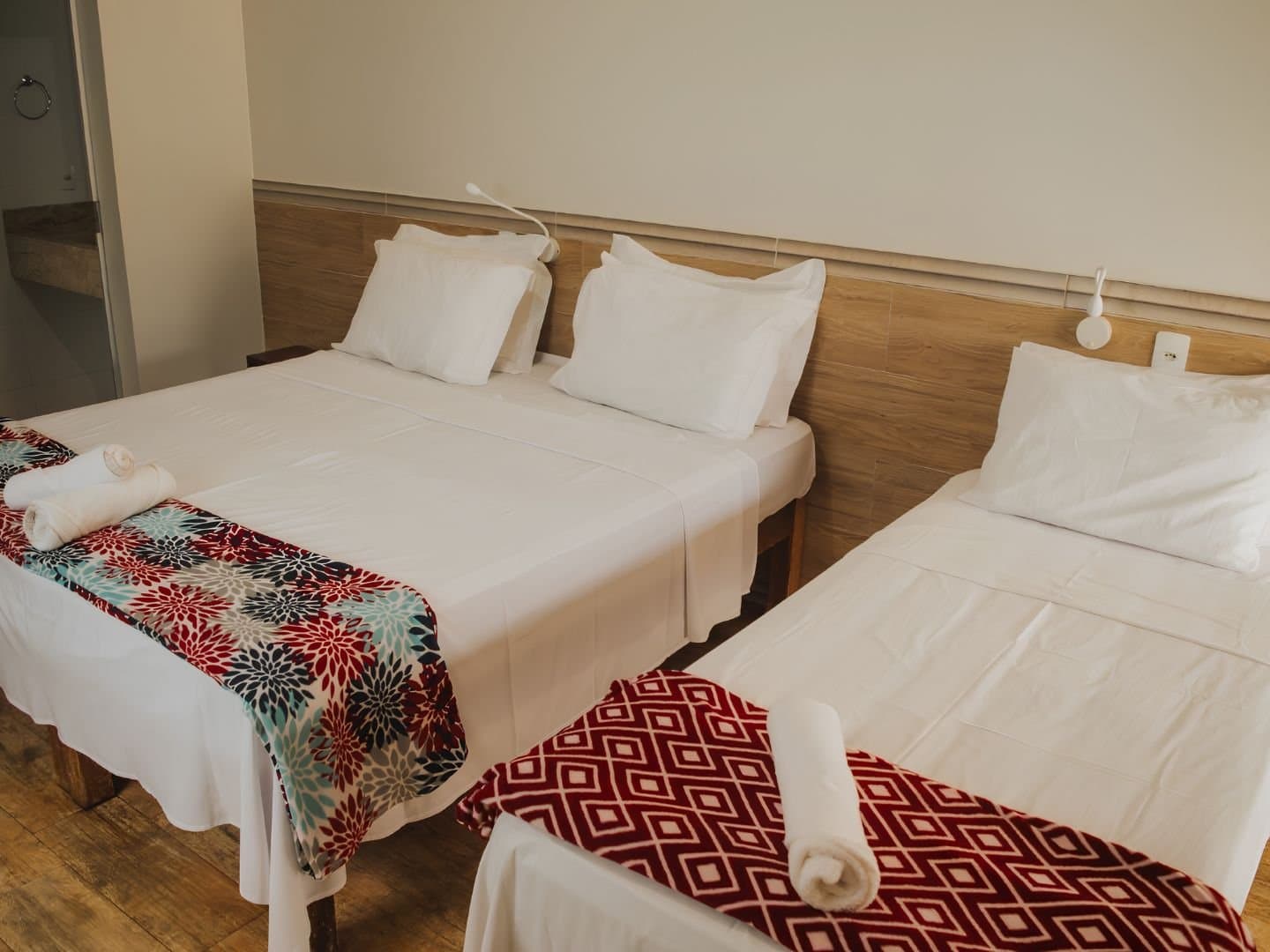 Cama arrumada, toalha enrolada, e o trilho de amanhã já esperando por você. Quarto triplo em pousada de aventura no Jalapão com duas camas de casal e uma de solteiro, roupa de cama branca, pés de cama floridos e geométricos e toalhas enroladas sobre piso de madeira
