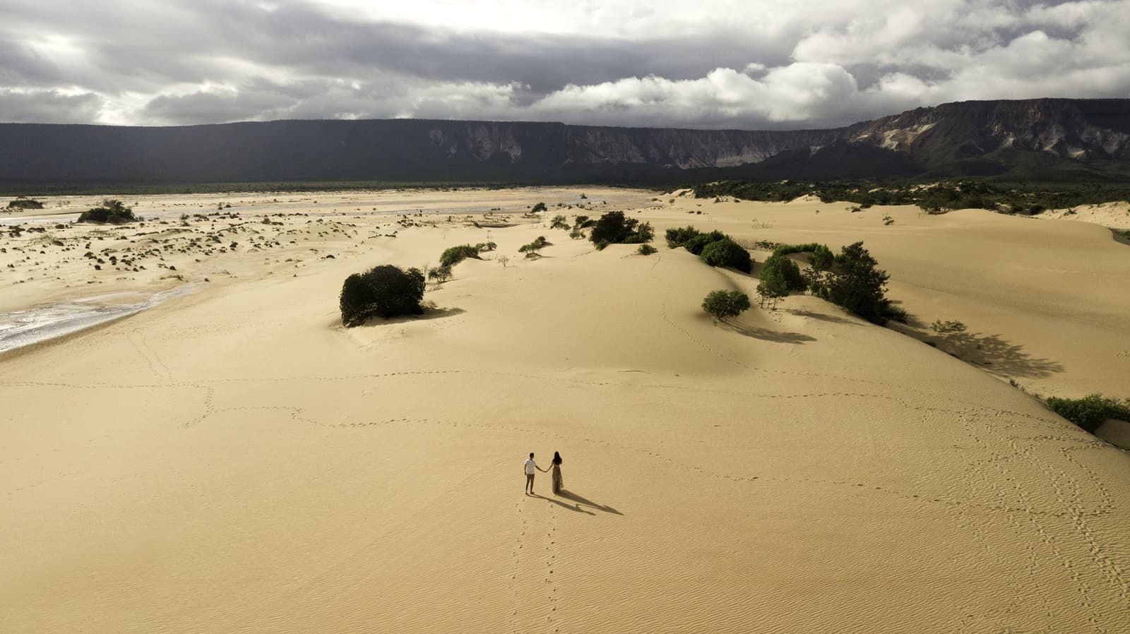 Zwei Menschen, eine riesige Düne, und Spuren, die der Wind morgen schon verwischt hat. Photo: FG Trade Luftaufnahme eines Pärchens, das sich an den goldenen Sanddünen von Jalapão an den Händen hält, mit grünen Sträuchern, einem Fluss und einer dramatischen Sandstein-Steilwand unter bewölktem Himmel