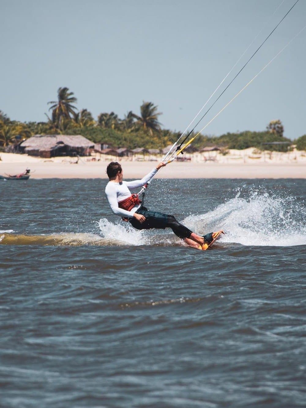 Der Wind wartet nicht, er überzeugt auf seine eigene Art. Ein Kitesurfer gleitet über aufgewühltes Wasser vor einem weißen Sandstrand mit Palmen und Strohhütten im Hintergrund