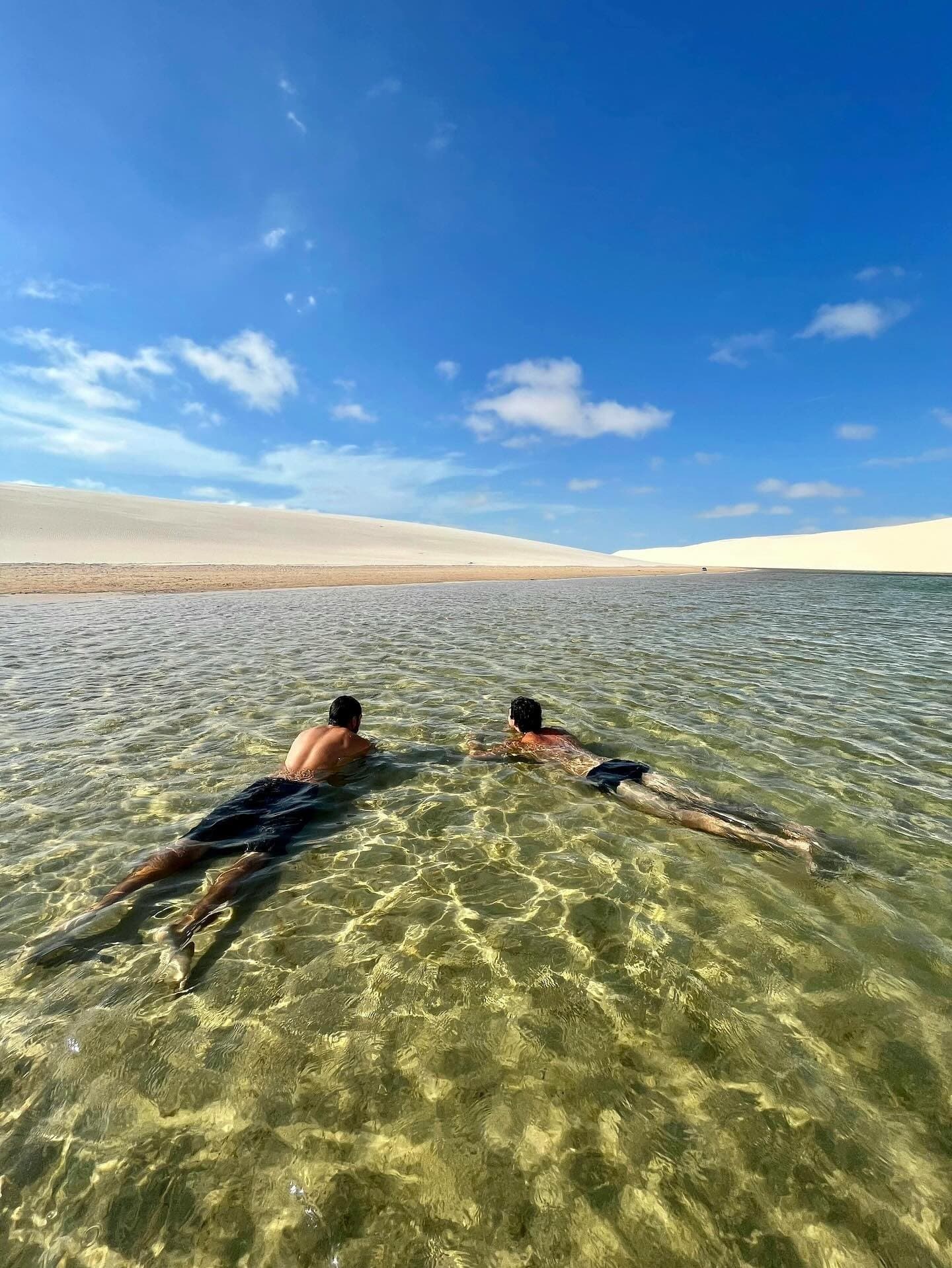 Das Wasser ist so klar, dass man jeden Sandkorn auf dem Grund sehen kann. Zwei Schwimmer treiben bäuchlings in einer kristallklaren, flachen Lagune in den Lençóis Maranhenses, im Hintergrund erstrecken sich weiße Sanddünen bis zum Horizont