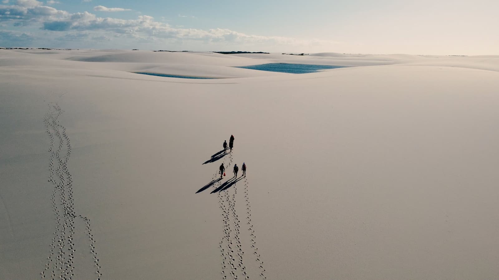 Vier Menschen, Sand ohne Ende und das gute Gefühl, mal wirklich winzig zu sein. Luftaufnahme von vier Wanderern, die auf der Lençóis-Maranhenses-Wanderung über weitläufige weiße Sanddünen laufen, mit langen Schatten, Fußspuren und zwei blauen Lagunen in der Ferne