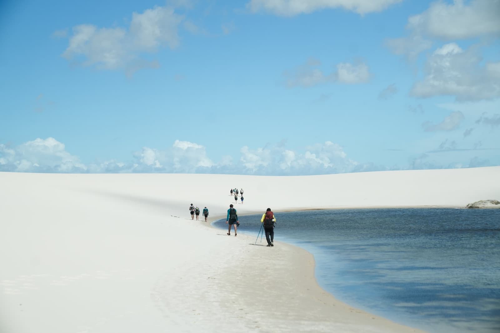 La piste tient en un fil de sable blanc entre lagune et dune. Un groupe de randonneurs avec des sacs à dos marchent en file indienne sur une langue de sable blanc entre une grande lagune bleue et des dunes immenses lors d'un trek dans les Lençóis Maranhenses