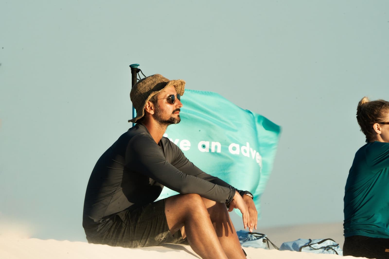 Un guide qui regarde au loin, et soudain vous regardez aussi. Un homme en chapeau de paille et lunettes de soleil est assis sur les dunes de sable blanc lors d'une pause du trekking des Lençóis Maranhenses, un drapeau turquoise claquant dans le vent derrière lui