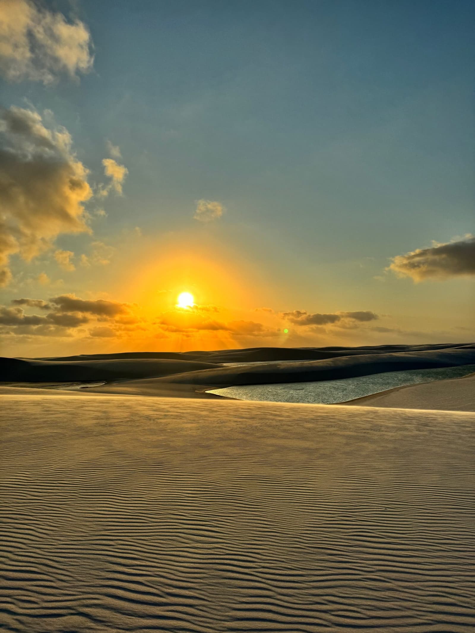 Un désert qui garde ses lagunes comme un secret bien gardé. Coucher de soleil doré sur des dunes de sable blanc ondulées avec une lagune peu profonde reflétant la lumière dans les Lençóis Maranhenses
