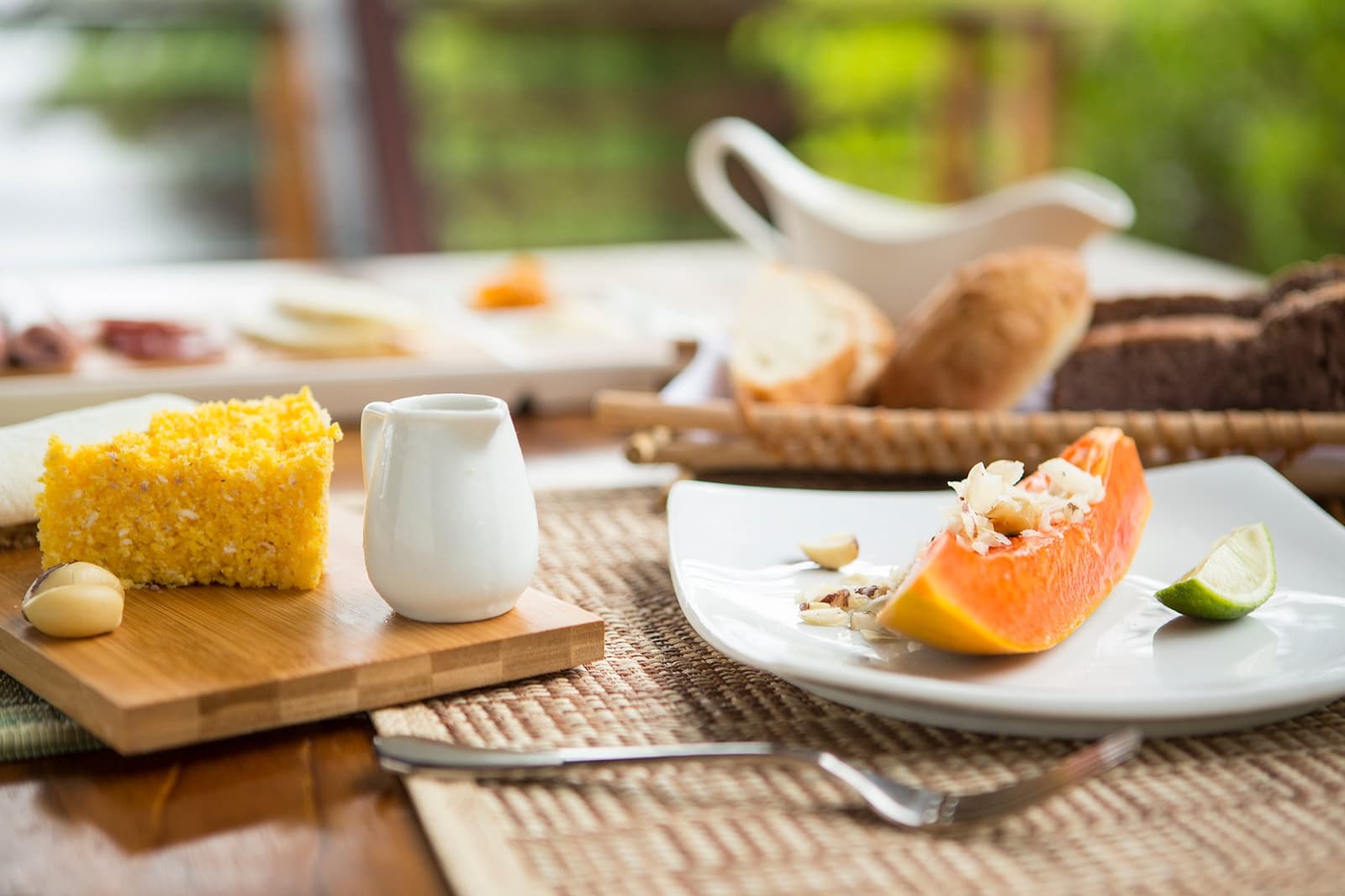 Mit reifer Papaya und hausgemachtem Kuchen in den Tag, der Regenwald läuft nicht weg. Ein frisches Papayastück mit Nüssen und Limette sowie ein Stück goldgelbes Maisbrot auf einem Holzbrett beim Frühstück im Mirante do Gavião, mit einem Brotkorb im Hintergrund