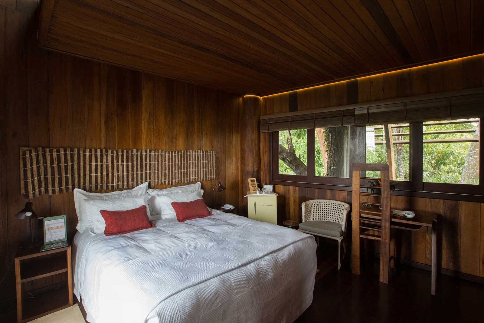 Holzgetäfelte Wände, Regenwald direkt vor dem Fenster, hier schläft man tief. Gästezimmer im Mirante do Gavião Amazon Lodge mit dunklen Holzwänden, weißer Bettwäsche, roten Dekokissen und Fenstern zum Regenwald