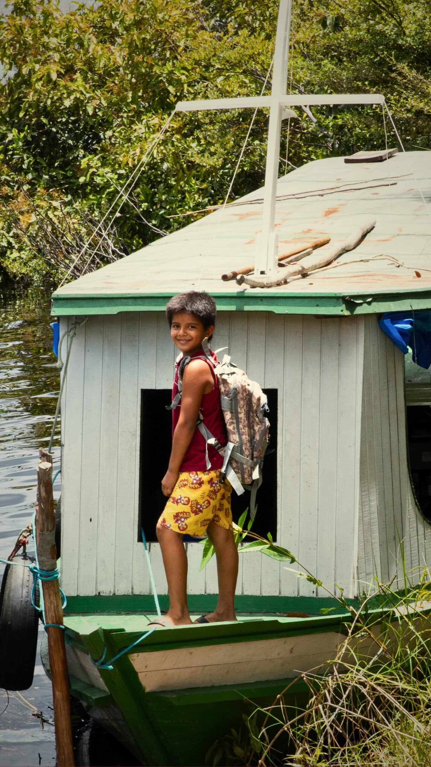 Schulweg auf Amazonisch: Rucksack drauf, Boot ablegen, fertig. Ein lächelnder Junge mit Rucksack steht barfuß auf einem kleinen Holzboot am Dschungelufer, das zu einer Flussgemeinde in der Nähe der Mirante do Gavião Lodge gehört