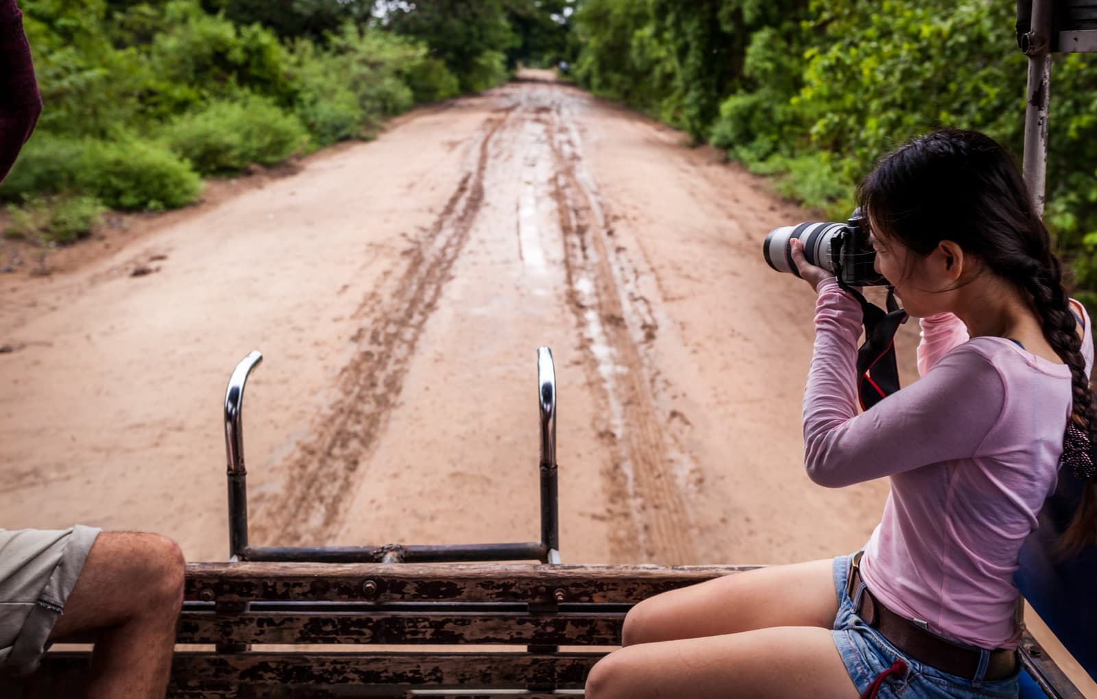 Camera up, the road ahead holds all the promise you need. Photo: nok6716 A young woman shoots with a telephoto lens from the back of a 4x4 vehicle on a muddy dirt road lined with green vegetation during a southern pantanal safari