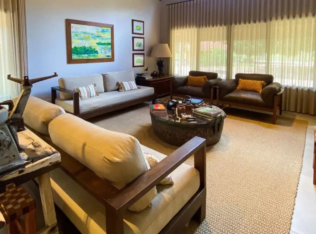 Good books, warm cushions, a coffee table full of field guides. Camp life has its perks. Comfortable lodge living room on a southern pantanal safari, featuring dark-wood sofas, a round antique coffee table stacked with wildlife books, colorful landscape paintings on the wall, and large curtained windows overlooking green vegetation