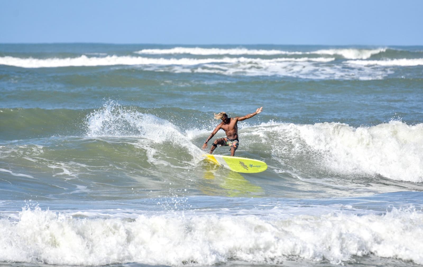 Braços abertos, cabelo solto, no ritmo certo. É assim que funciona. Photo: Picasa Um surfista de cabelos longos e loiros surfa numa prancha longboard amarela em uma onda quebrando durante aulas de surf em Itacaré, braços abertos para equilibrar sob um céu azul.