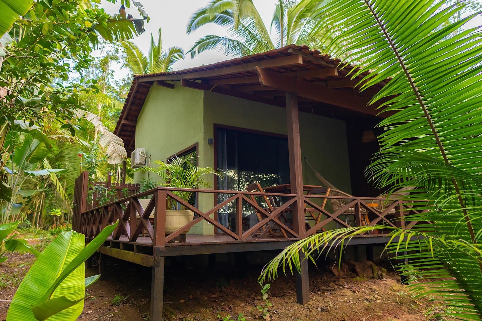 Wenn der Regenwald direkt vor der Terrassentür steht, ist das kein Zufall. Bungalow einer Amazon-Boutique-Lodge mit grüner Fassade, erhöhter Holzterrasse und Kreuzgeländer, eingebettet zwischen Palmwedeln, Bananenblättern und tropischem Blätterdach
