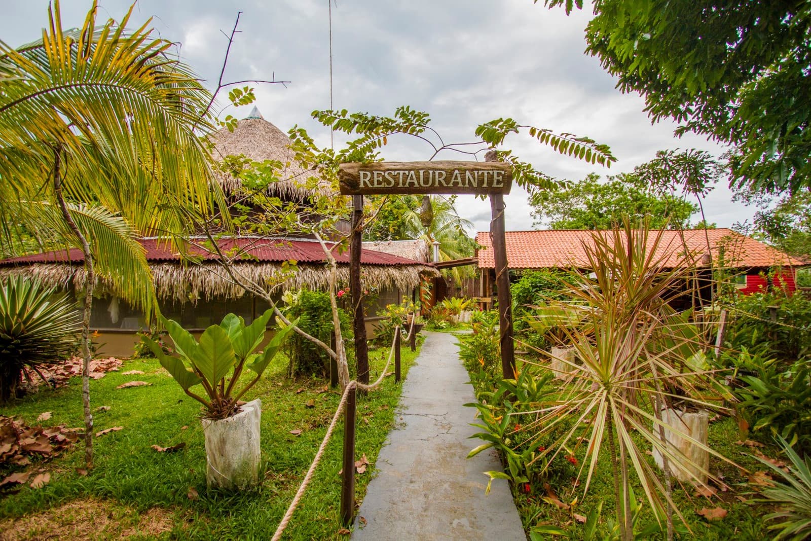 Das Mittagessen wartet am Ende des Wegs, der Dschungel ist schon nebenan. Ein hölzernes Schild mit der Aufschrift 'Restaurante' überspannt einen Gartenweg, der zu strohgedeckten und ziegelgedeckten Gebäuden des Amazon Turtle Lodge führt, umgeben von Palmen und üppigem Tropengrün