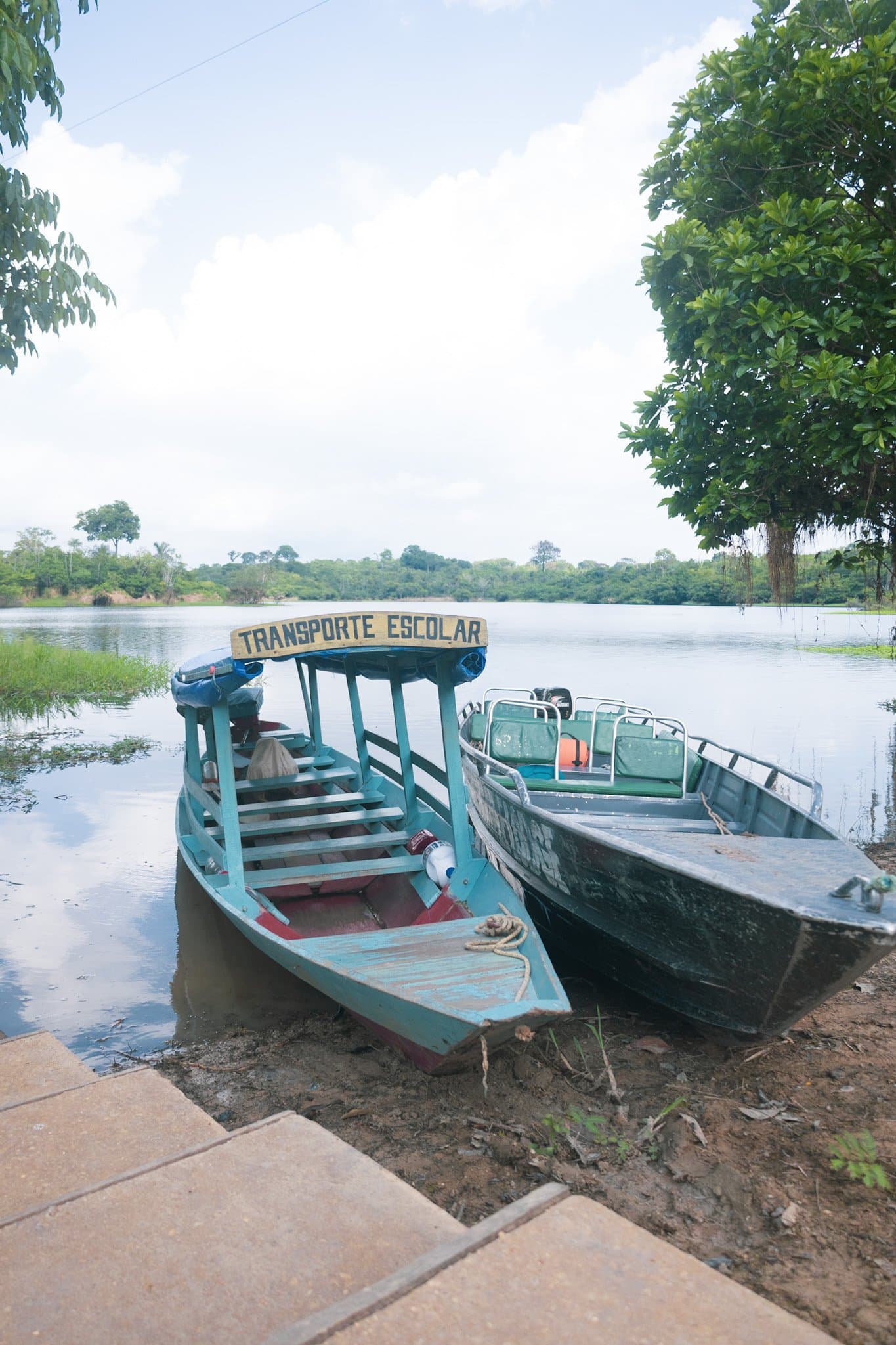 Schulbus? Hier ist es ein Schulboot, und das ist keine Metapher. Zwei hölzerne Motorboote am Ufer eines ruhigen Flusses nahe der Amazon Turtle Lodge, eines türkis mit dem Schriftzug 'Transporte Escolar', üppiger Regenwald spiegelt sich im stillen Wasser