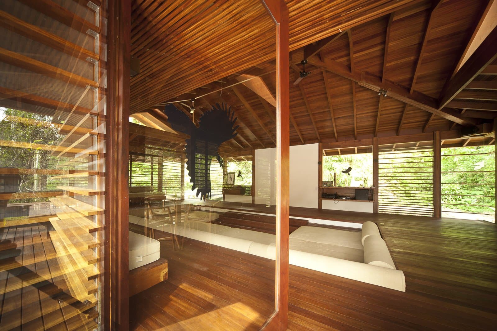 Ici, la forêt n'est pas le décor, elle est la pièce. Photo: Samuel Melim (c) Salle commune ouverte d'un lodge d'observation des oiseaux en forêt amazonienne, avec un plafond voûté en bois dur, des banquettes blanches intégrées, des volets en bois à lamelles et une grande sculpture d'oiseau suspendue aux chevrons, la forêt verte luxuriante visible de tous côtés