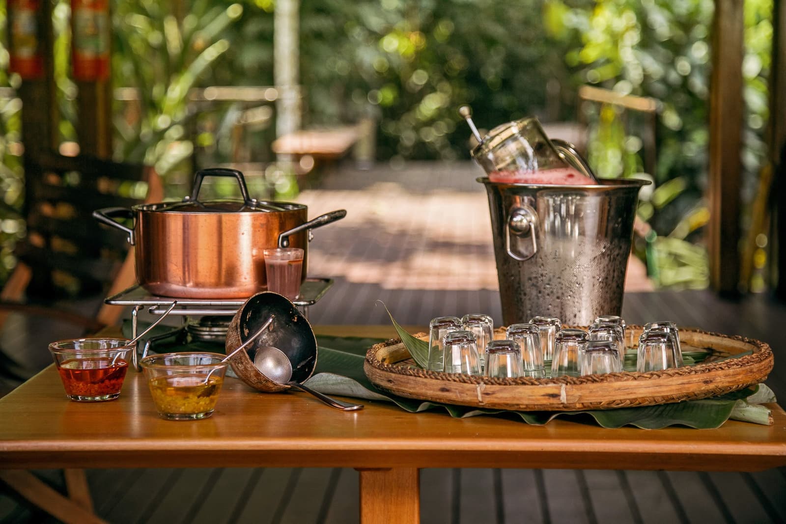Le pot chauffe, les verres attendent. Bienvenue, à la manière de l'Amazonie. Photo: | SamuelMelim Photography Casserole en cuivre chauffant sur un réchaud portable à côté d'un seau à glace martelé rempli d'un jus rose et de bocaux en verre, petits verres à shot disposés sur un plateau tressé tapissé de feuilles de bananier, et deux bols de condiments sur une table en bois d'une terrasse de lodge en plein air