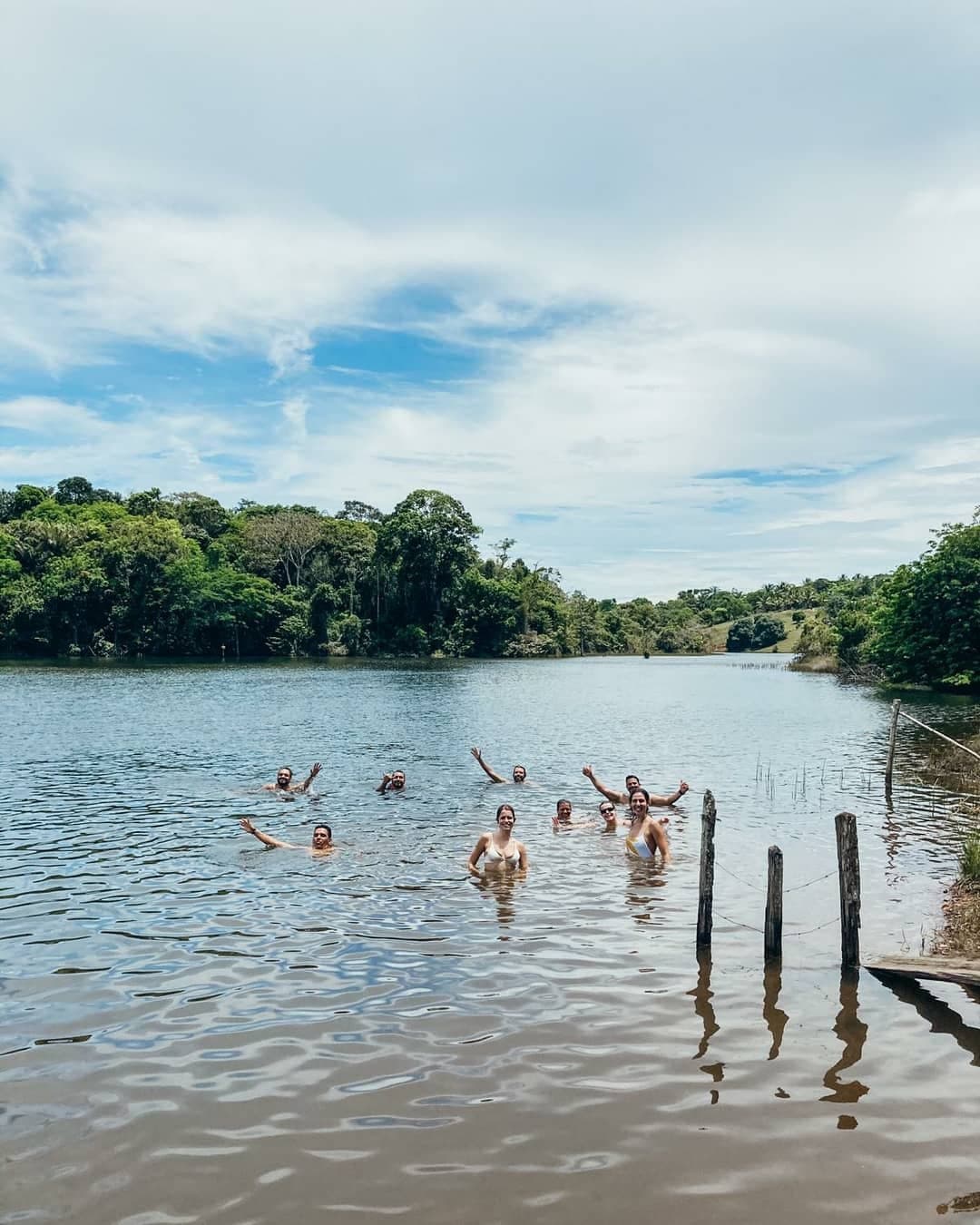 Mittagspause verdient, Räder geparkt, alle gleichzeitig ins Wasser. Eine Gruppe von Radreisenden schwimmt in einem ruhigen See, umgeben von dichtem grünen Wald in Bahia, und winkt in die Kamera