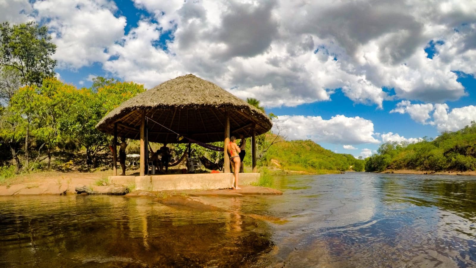 Hängematte, Schatten, Fluss. Mehr braucht es nach einem langen Wandertag nicht. Ein strohgedecktes Freiluftpavillon mit Hängematten steht auf einer niedrigen Steinplattform an einem klaren, flachen Fluss im Jalapão, mit grünen Cerrado-Hügeln und einem dramatischen Wolkenhimmel; eine Reisende im Bikini steht am Rand und blickt über das Wasser