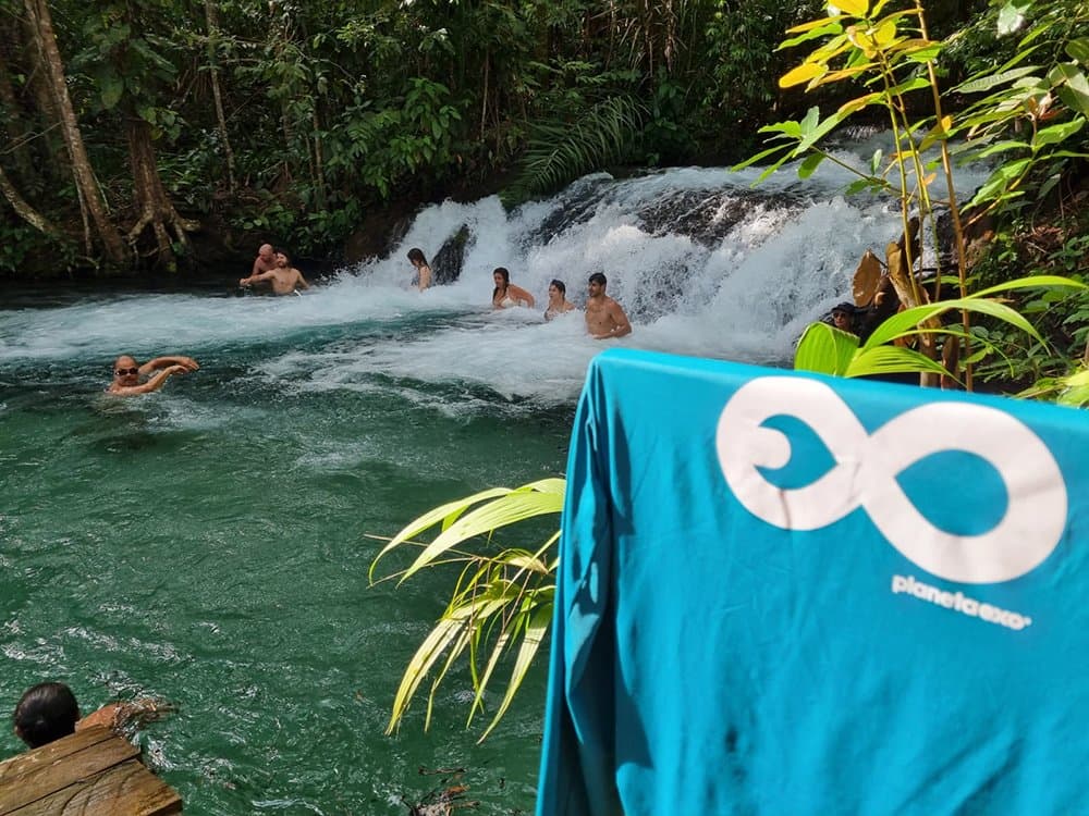 Kaltes Wasser, dichtes Blätterdach, und kein vernünftiger Grund, da rauszukommen. Eine Gruppe von Reisenden schwimmt und watet an der Basis eines rauschenden Wasserfalls, umgeben von dichtem tropischem Regenwald, beim Jalapão-Trekking, mit einem PlanetaEXO-Handtuch im Vordergrund