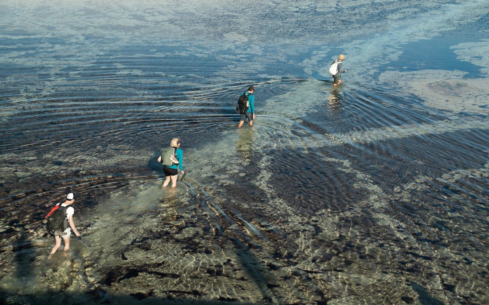 Nasse Füße, klares Wasser, kein Bedauern. Vier Wanderer durchwaten flaches, kristallklares Wasser auf einer Wanderung durch die Lençóis Maranhenses, ihre Wellen breiten sich über einen felsigen Flussboden aus, von oben fotografiert