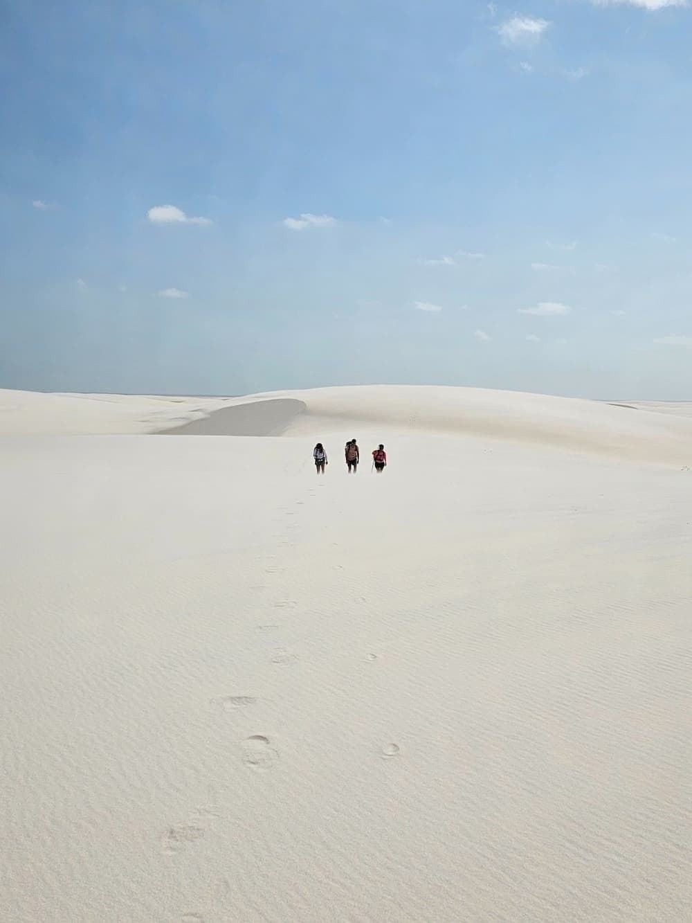 Drei Menschen, endloser Sand, keine Eile. Drei Wanderer mit Rucksäcken überqueren die weiten weißen Sanddünen des Lençóis Maranhenses, ihre Fußspuren im Sand unter einem hellblauen Himmel