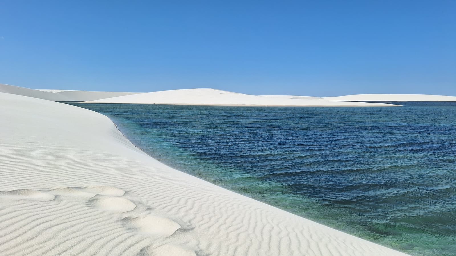 Kaltes Wasser, heißer Sand, ein Kontrast, der sich nie abnutzt. Gerippelte weiße Sanddünen treffen auf eine breite blau-grüne Süßwasserlagune unter klarem blauen Himmel beim Lençóis-Maranhenses-Trekking, mit weiteren Dünen am gegenüberliegenden Ufer