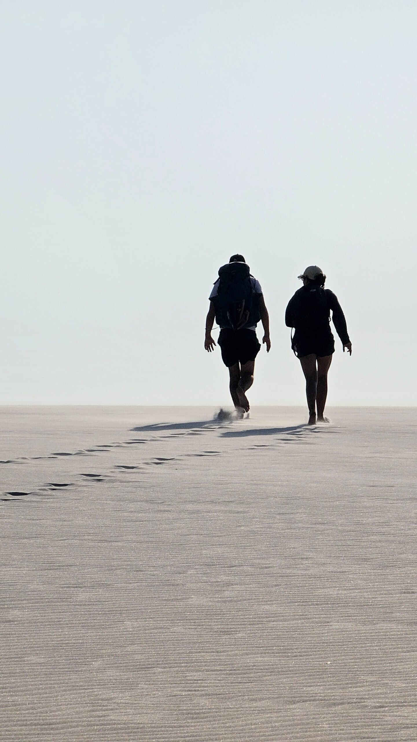 Weißer Sand bis zum Horizont, und ihr seid die Einzigen, die ihn durchqueren. Zwei Wanderer laufen auf dem Lençóis-Maranhenses-Trek von Atins über eine weite, flache Weißsandfläche, ihre Fußspuren verlieren sich im dunstigen Horizont