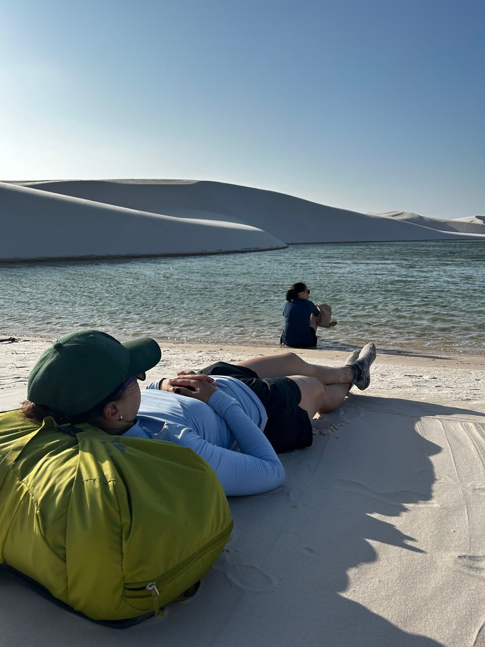 Les dunes peuvent attendre. Certains moments se vivent à plat dos. Un randonneur allongé sur le sable blanc d'une lagune limpide lors d'un trek dans les Lençóis Maranhenses, une deuxième personne assise au bord de l'eau devant de grandes dunes blanches