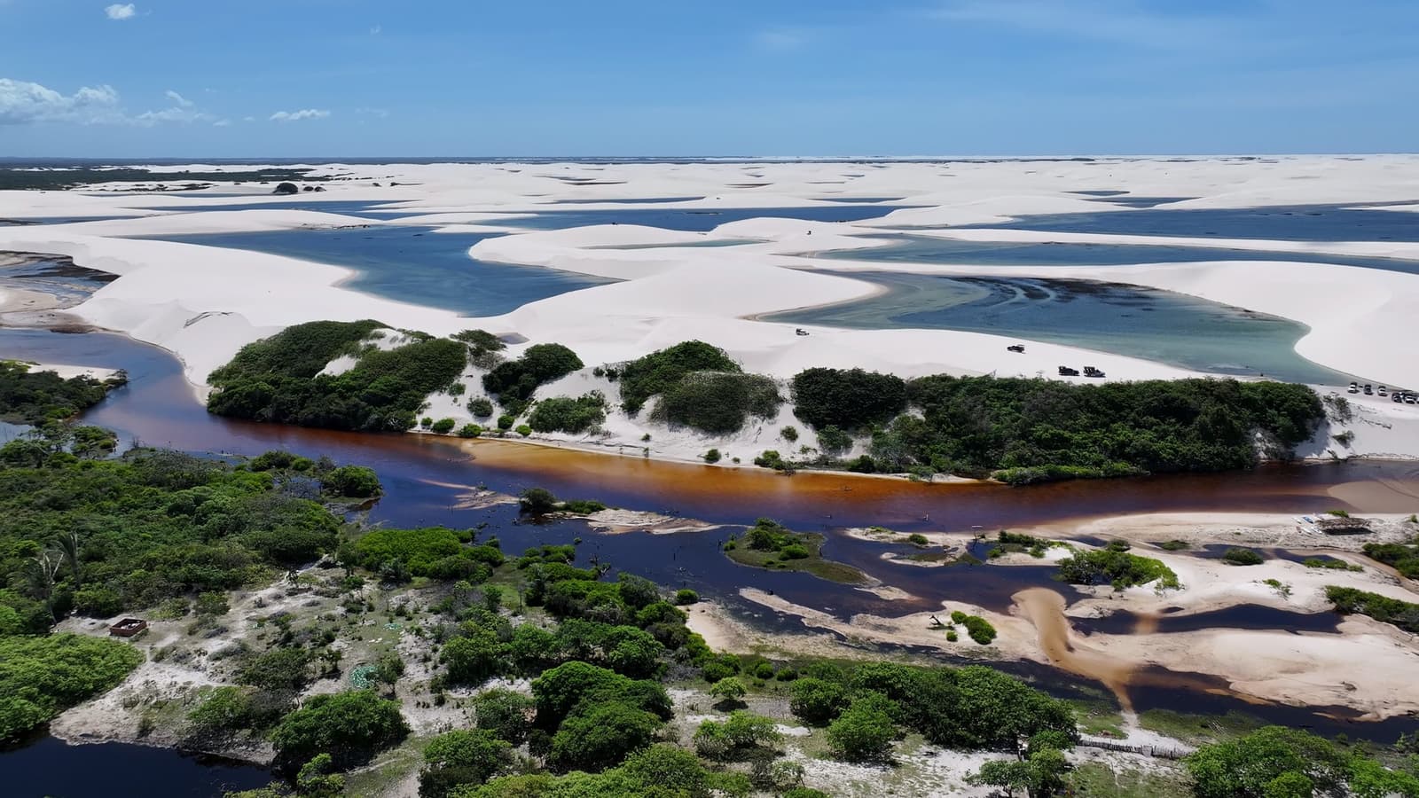 Une rivière noire, des dunes blanches, des lagunes bleues. La géographie ici refuse de choisir. Vue aérienne d'une rivière aux eaux tanniques serpentant à travers la végétation avant de rejoindre les dunes de sable blanc et les lagunes turquoise du trek des Lençóis Maranhenses