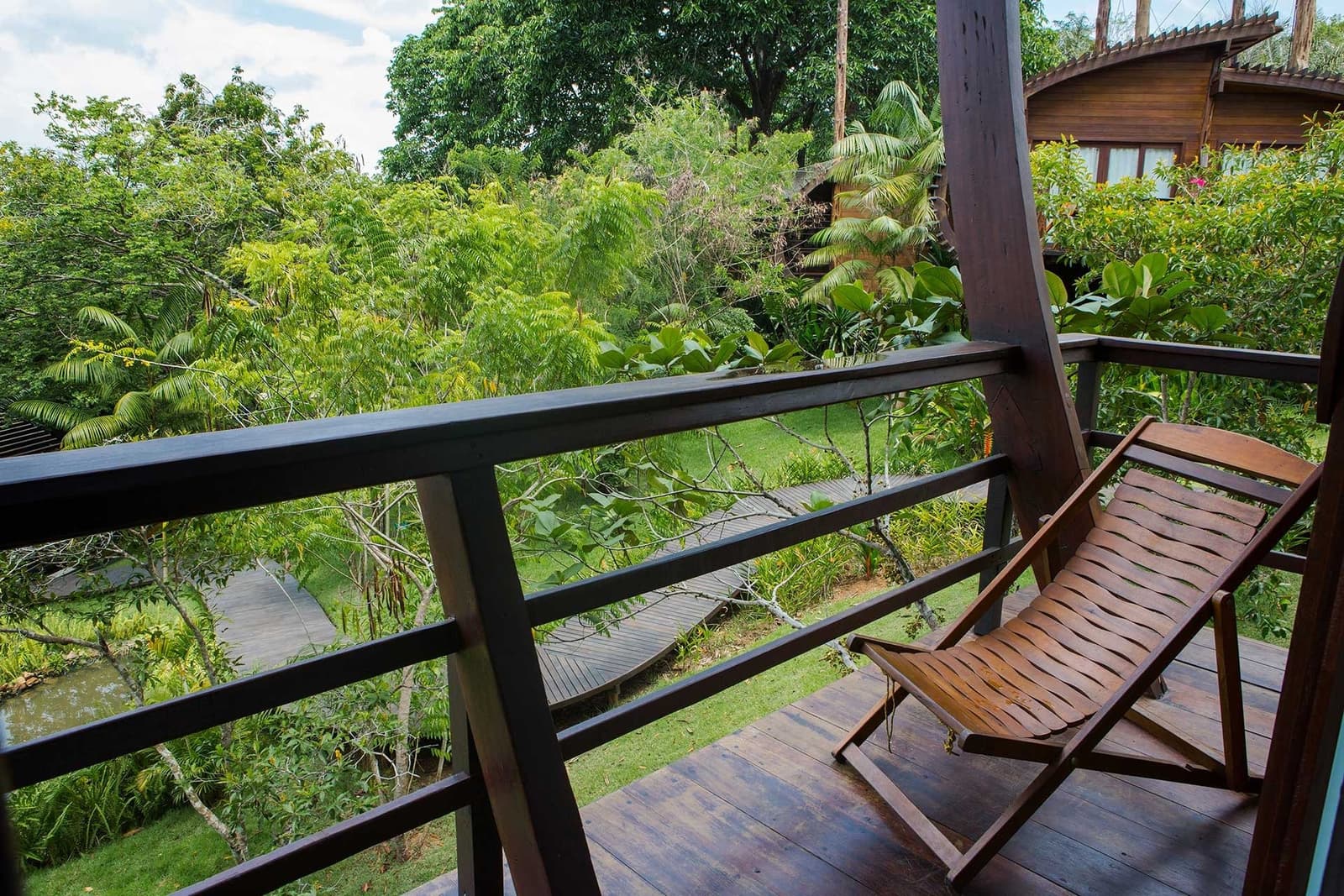 Hier sitzt man einfach und schaut, wie der Dschungel atmet. Ein Holzliegestuhl auf dem Holzbalkon des Mirante do Gavião mit Blick auf einen üppigen tropischen Garten, Holzsteg und benachbartem Bungalow