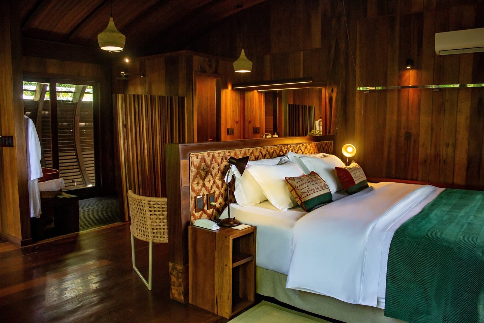 Nach einem Tag auf dem Fluss weiß man dieses Zimmer wirklich zu schätzen. Suite im Mirante do Gavião Amazon Lodge mit Kingsize-Bett in weißer Bettwäsche, handgewebten Kissen, Rattankopfteil, warmen Pendelleuchten und dunklen Holzwänden mit Blick auf ein naturnahes Außenbad