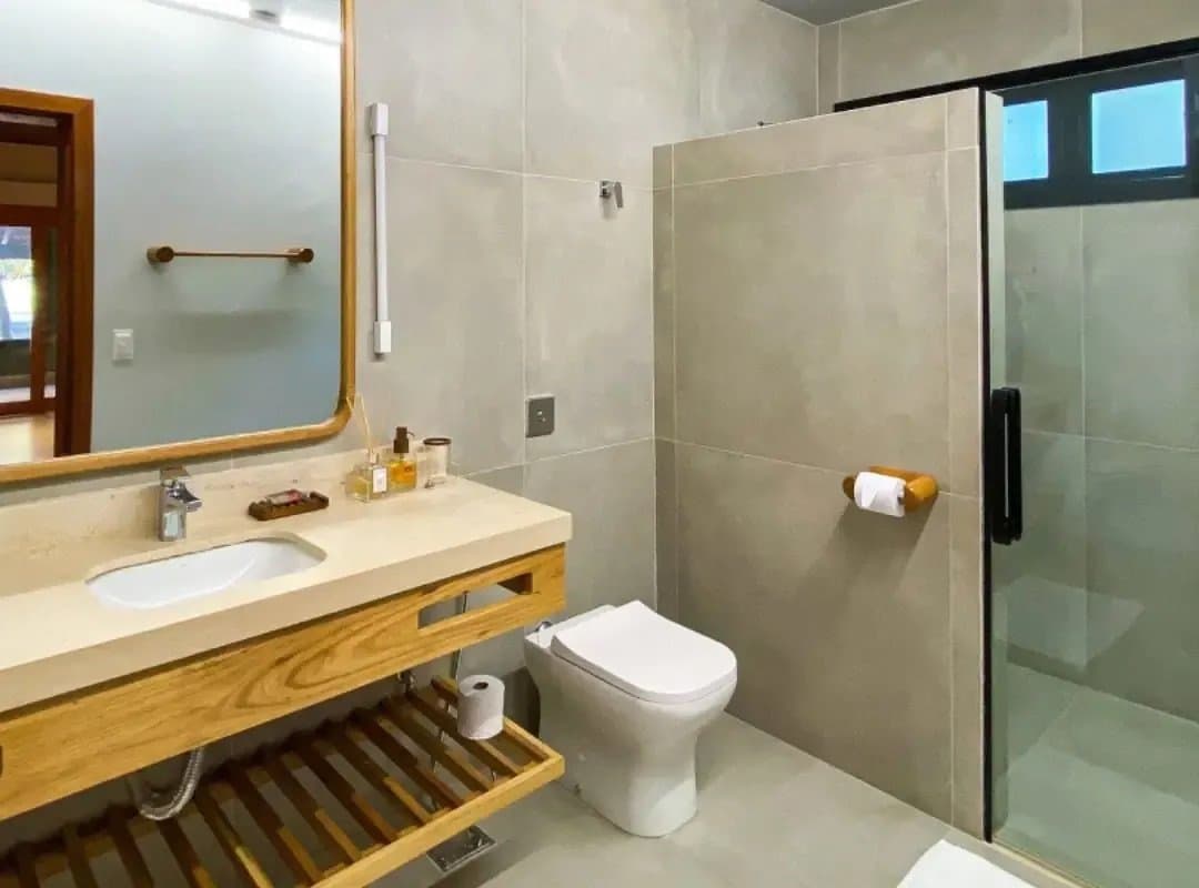 Hot water, wood accents, proper toiletries. Comfort doesn't stop at the forest edge. Clean modern bathroom at a southern pantanal safari lodge, featuring a warm wood vanity, stone countertop, large mirror, and glass-enclosed shower with neutral tile walls