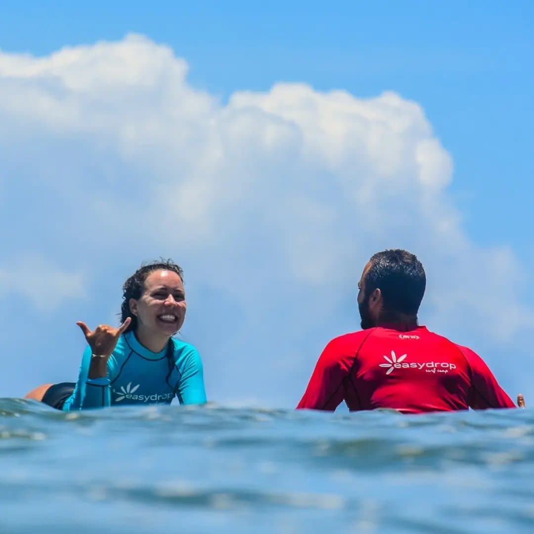 Um shaka que dispensa qualquer nota. Uma aluna sorridente com lycra EasyDrop azul-turquesa faz o sinal de shaka para seu instrutor durante uma aula de surf em Itacaré, ambos sentados nas pranchas no mar aberto sob céu azul.