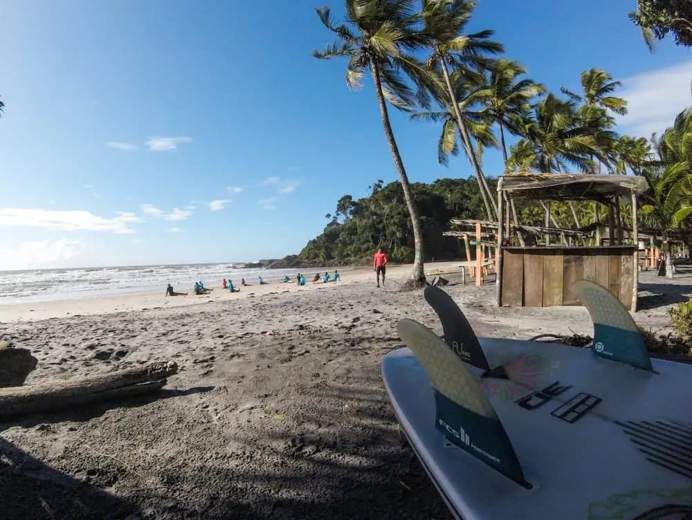 Boards in het zand, instructeur klaar, de dag kan beginnen. Een surfboard met FCS-vinnen ligt op het zand bij een surfschool in Itacaré, terwijl een groep leerlingen oefent aan de waterlijn voor palmbomen en Atlantisch regenwoud.